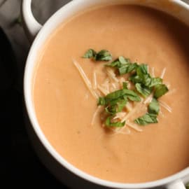 A bowl of creamy tomato soup garnished with grated cheese and fresh basil leaves.