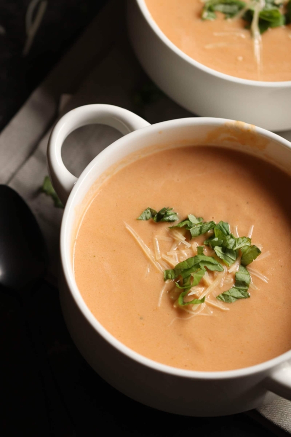 A bowl of creamy tomato soup garnished with grated cheese and fresh basil leaves.