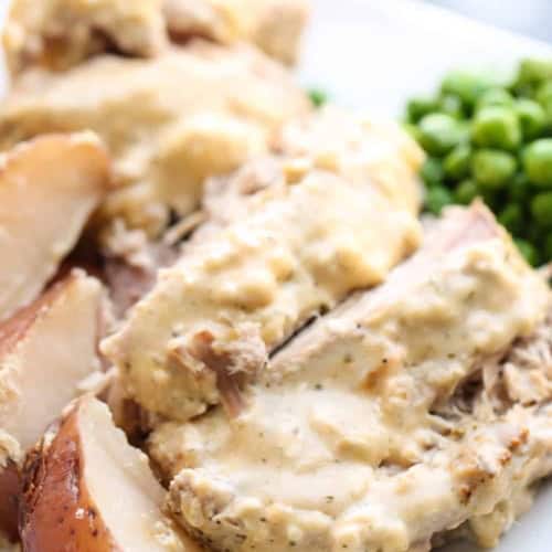 Slices of pork roast with creamy gravy, served with red potatoes and green peas on a white plate.