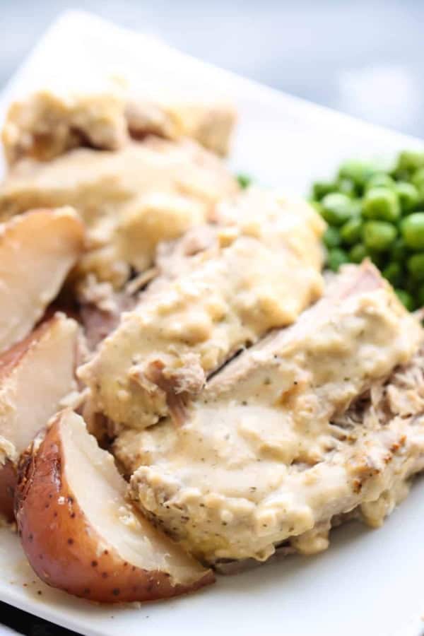 Slices of pork roast with creamy gravy, served with red potatoes and green peas on a white plate.