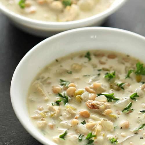 Two white bowls filled with creamy chili containing beans, chicken, herbs, and garnished with chopped parsley.