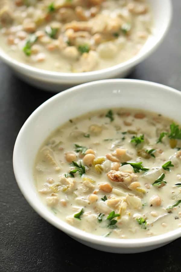 Two white bowls filled with creamy chili containing beans, chicken, herbs, and garnished with chopped parsley.