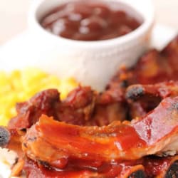 A plate of barbecue pork ribs with a side of yellow rice and a cup of barbecue sauce.