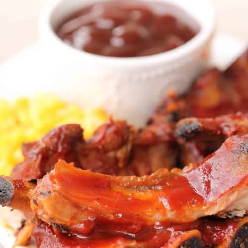 A plate of barbecue pork ribs with a side of yellow rice and a cup of barbecue sauce.