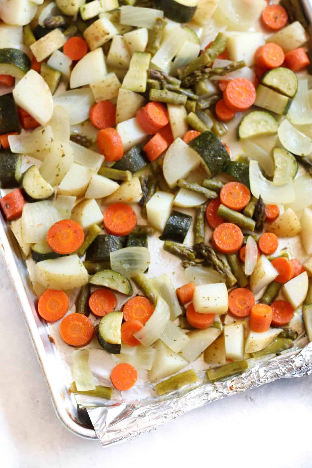 A baking sheet lined with foil holds roasted vegetables including carrots, zucchini, onions, potatoes, and asparagus.