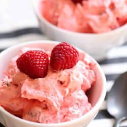 Two white bowls filled with pink creamy dessert topped with fresh raspberries, placed on a striped cloth next to a spoon.