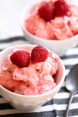 Two white bowls filled with pink creamy dessert topped with fresh raspberries, placed on a striped cloth next to a spoon.