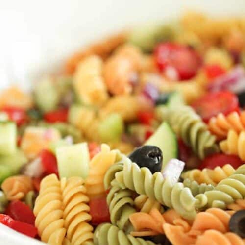 A close-up of a colorful pasta salad with tri-color rotini, sliced black olives, chopped cucumber, red onion, and red bell peppers in a white bowl.