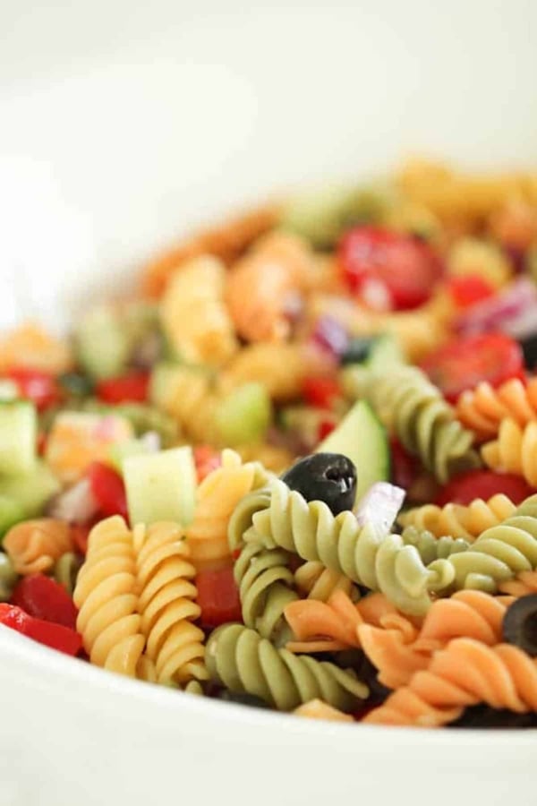 A close-up of a colorful pasta salad with tri-color rotini, sliced black olives, chopped cucumber, red onion, and red bell peppers in a white bowl.