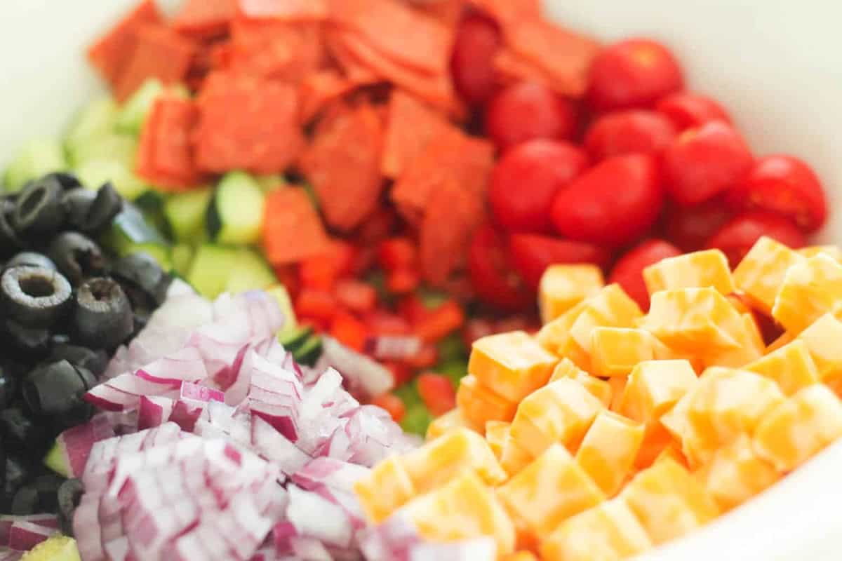 A bowl with chopped red onions, black olives, cubed cheese, cherry tomatoes, diced peppers, cucumbers, and sliced pepperoni.