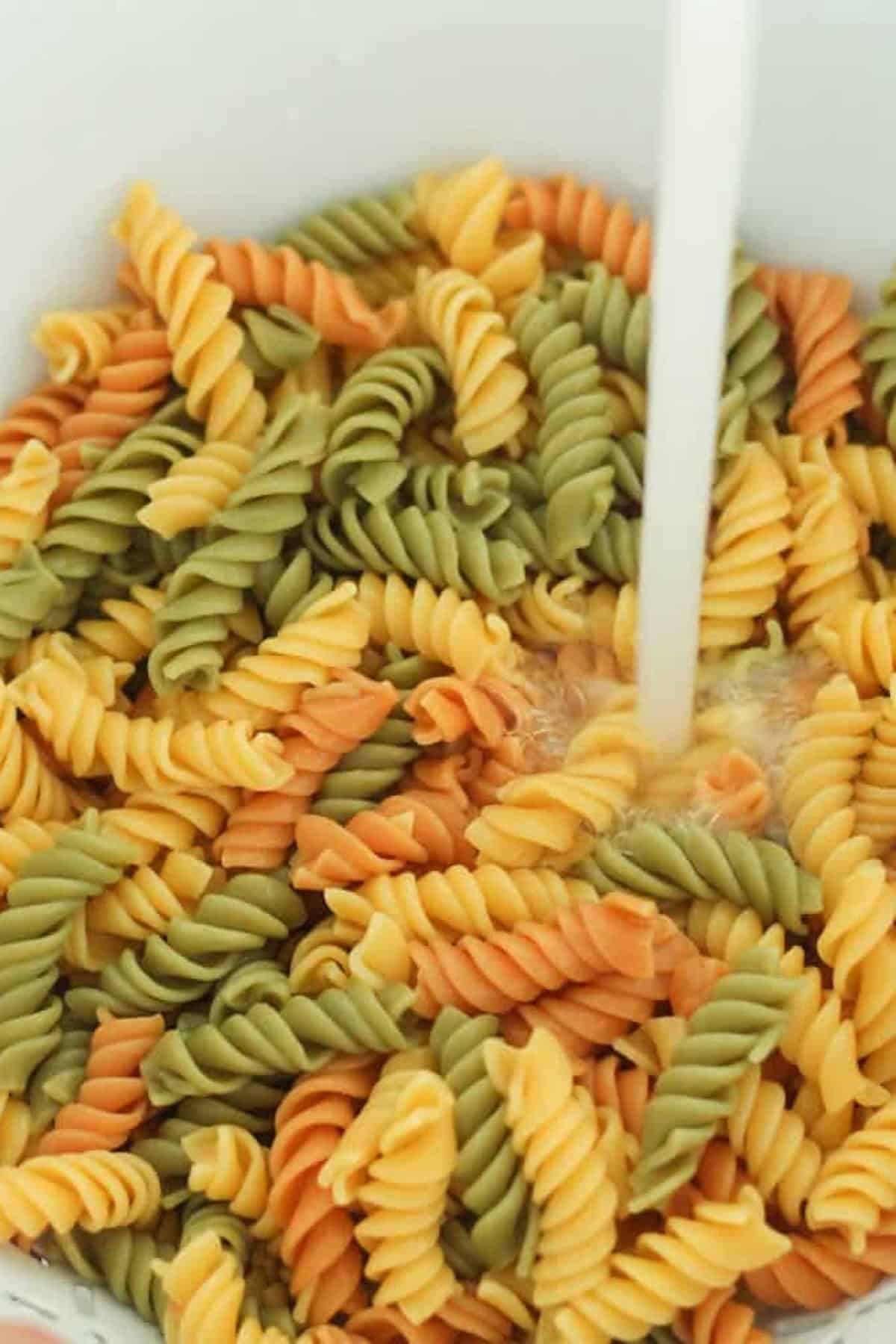 Tri-color rotini pasta being rinsed under running water in a colander.