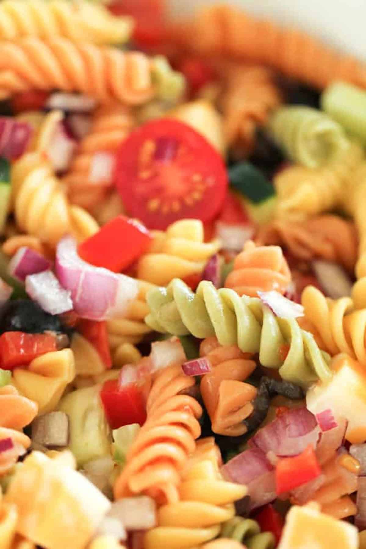 Close-up of a colorful pasta salad with rotini noodles, cherry tomatoes, diced red and green bell peppers, red onions, and sliced olives.