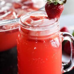 A mason jar filled with bright red strawberry punch, ice cubes, and a whole strawberry garnish, with a bowl of punch in the background.