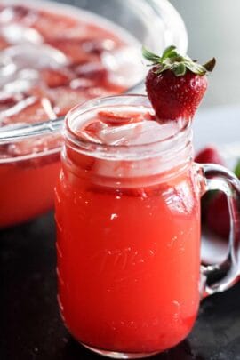 A mason jar filled with bright red strawberry punch, ice cubes, and a whole strawberry garnish, with a bowl of punch in the background.