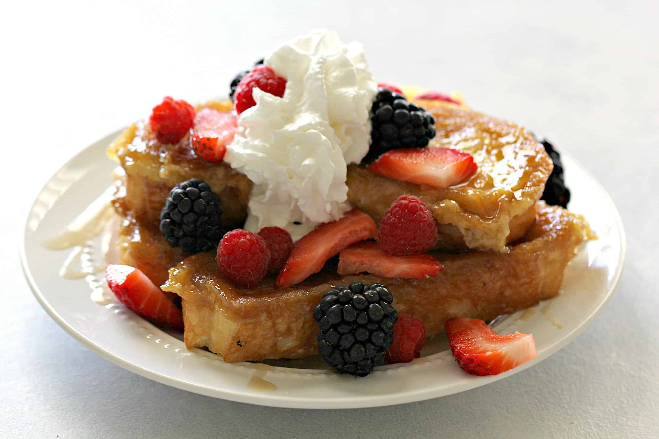 stack of creme brulee french toast topped with raspberries, strawberries, blackberries, and whipped cream