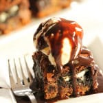 Ultimate Chocolate Chip Cookie n' Oreo Fudge Brownie Bar topped with ice cream and hot fudge on a plate.