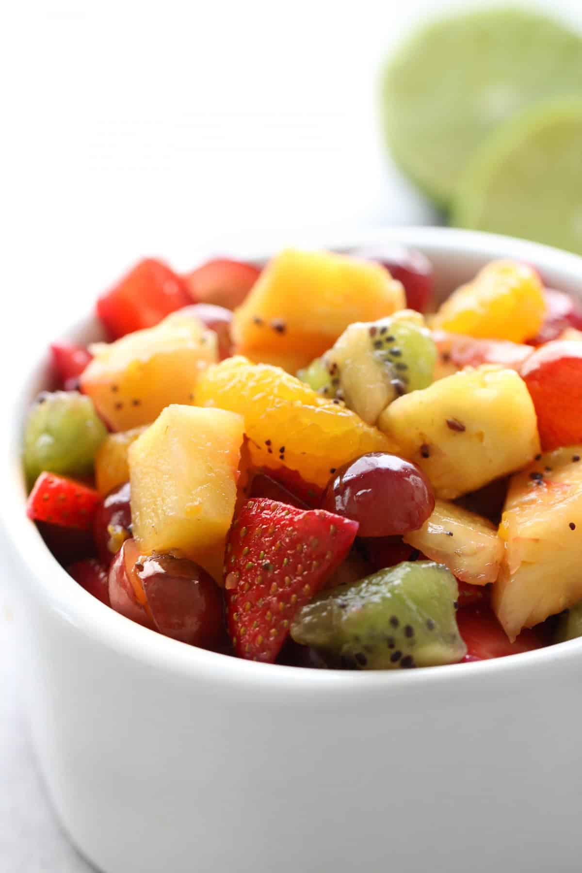 A white bowl filled with a colorful fruit salad containing pineapple, strawberries, grapes, kiwi, and orange pieces.