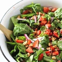 A bowl of fresh spinach salad with sliced strawberries, red onions, chopped walnuts, and a wooden serving spoon.