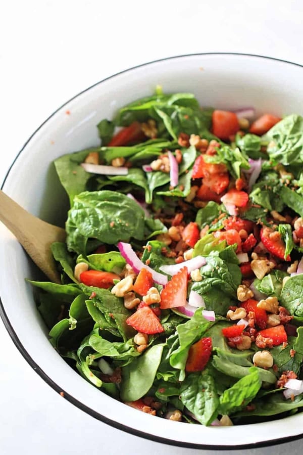 A bowl of fresh spinach salad with sliced strawberries, red onions, chopped walnuts, and a wooden serving spoon.