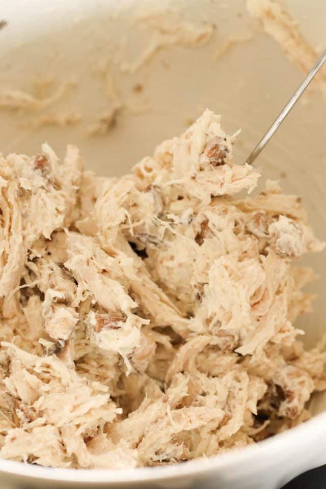 A bowl containing shredded chicken mixed with small pieces of cheese ingredients, stirred with a stainless steel fork.