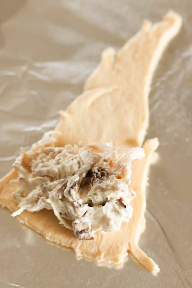 A triangular piece of dough is laid on a surface with a small amount of creamy mixture containing pieces of mushroom placed in the center.