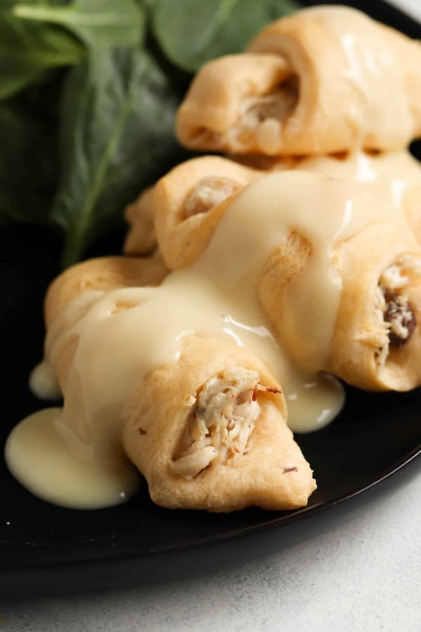Close-up of stuffed pastry rolls drizzled with a creamy white sauce on a black plate, accompanied by fresh spinach leaves.