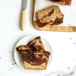 Two slices of a marbled loaf cake on a white plate, with more slices and a knife on a wooden cutting board nearby. Crumbs are scattered on the white surface.