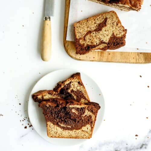 Two slices of a marbled loaf cake on a white plate, with more slices and a knife on a wooden cutting board nearby. Crumbs are scattered on the white surface.