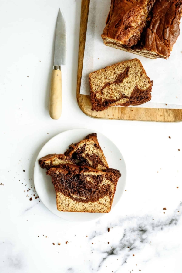 Two slices of a marbled loaf cake on a white plate, with more slices and a knife on a wooden cutting board nearby. Crumbs are scattered on the white surface.
