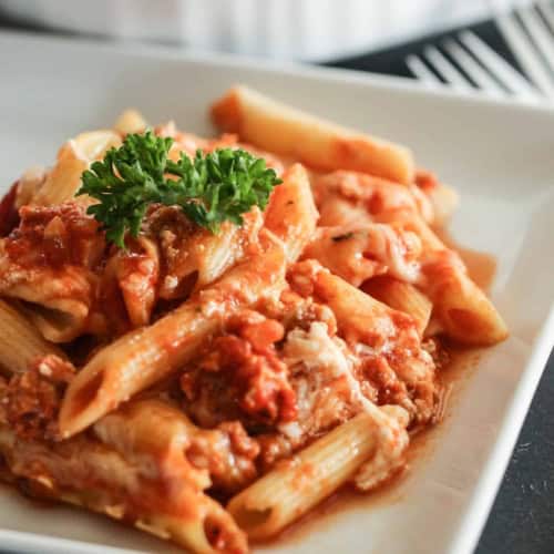 A serving of baked penne pasta with tomato sauce, ground meat, and melted cheese, garnished with a sprig of parsley on a white plate.