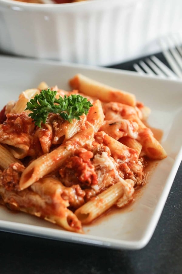 A serving of baked penne pasta with tomato sauce, ground meat, and melted cheese, garnished with a sprig of parsley on a white plate.