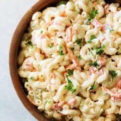 A wooden bowl filled with creamy macaroni salad garnished with chopped parsley. The salad includes elbow pasta, shredded carrots, and small pieces of vegetables.