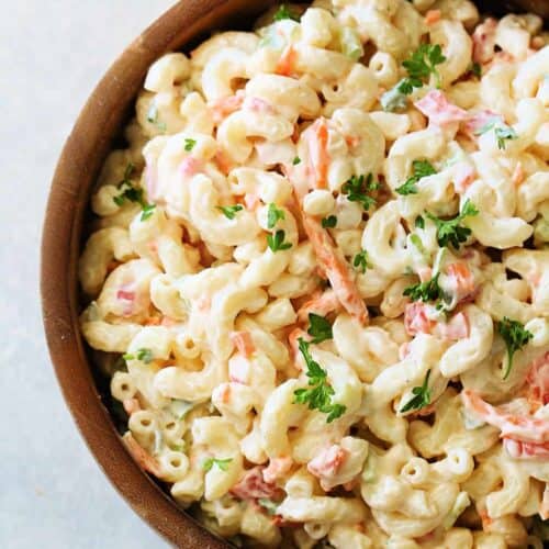 A wooden bowl filled with creamy macaroni salad garnished with chopped parsley. The salad includes elbow pasta, shredded carrots, and small pieces of vegetables.