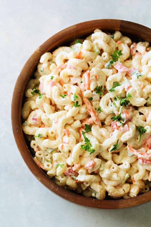 A wooden bowl filled with creamy macaroni salad garnished with chopped parsley. The salad includes elbow pasta, shredded carrots, and small pieces of vegetables.