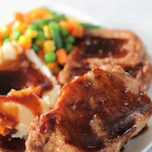 A plate with pork chops covered in brown gravy, mashed potatoes with gravy, and a side of mixed vegetables including green beans, corn, and carrots.