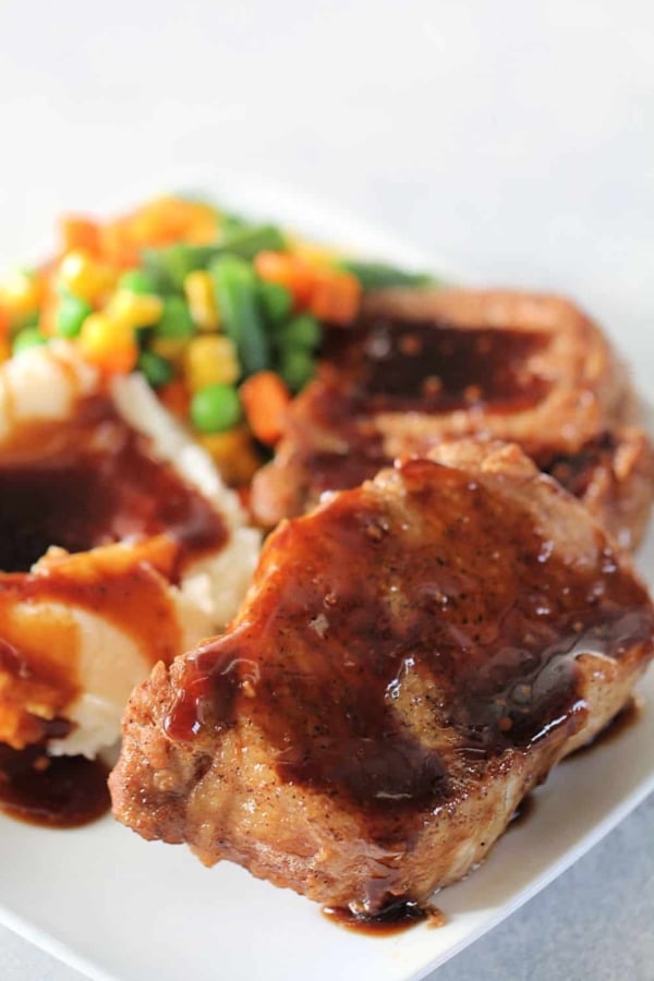 A plate with pork chops covered in brown gravy, mashed potatoes with gravy, and a side of mixed vegetables including green beans, corn, and carrots.