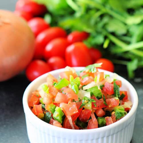 A small white bowl with Fresh Tomato Salsa. Fresh tomatoes and cilantro in the background.