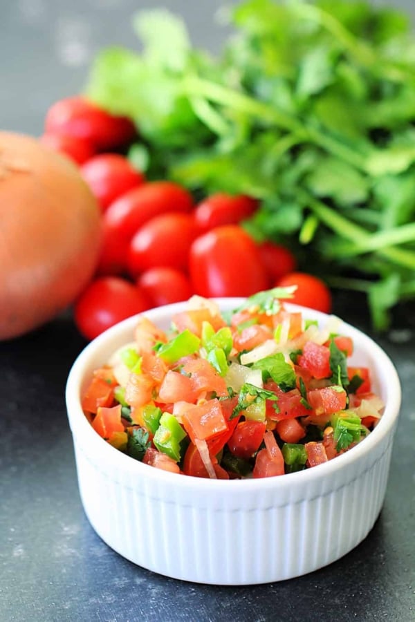 A small white bowl with Fresh Tomato Salsa. Fresh tomatoes and cilantro in the background.