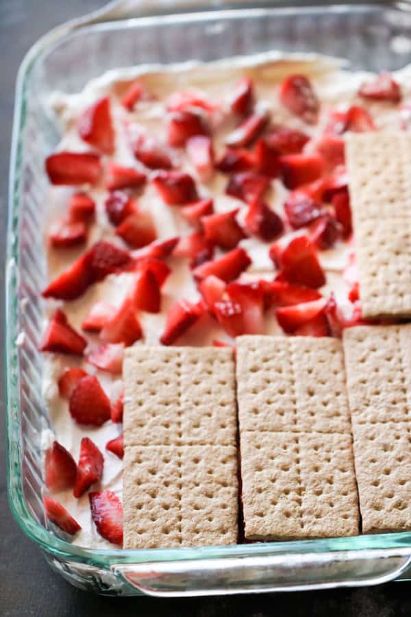 Sliced strawberries and cream in a baking dish with a layer of graham crackers being added on top.