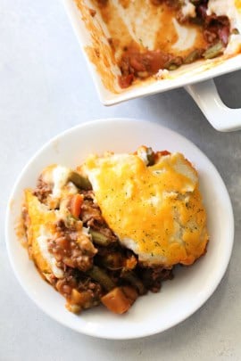 A white plate contains a serving of shepherd's pie topped with melted cheese, with visible ground meat, vegetables, and mashed potatoes. In the background, the dish it was served from is partially visible.
