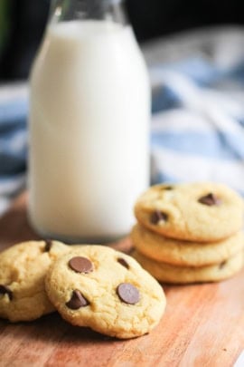 A bottle of milk stands next to a stack of three chocolate chip cookies with additional cookies scattered nearby on a wooden surface.