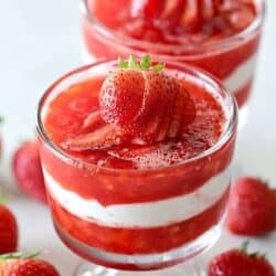 Two glass cups filled with layered strawberry and cream dessert, topped with sliced strawberries, surrounded by whole strawberries on a white surface.