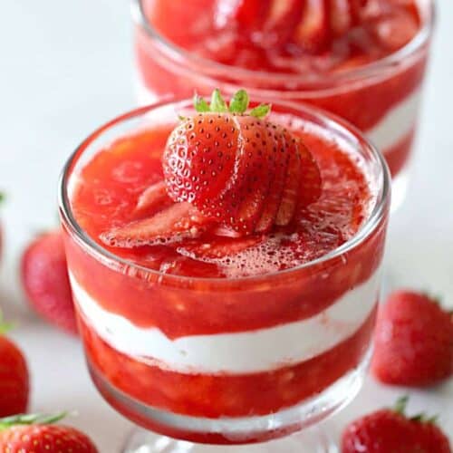 Two glass cups filled with layered strawberry and cream dessert, topped with sliced strawberries, surrounded by whole strawberries on a white surface.
