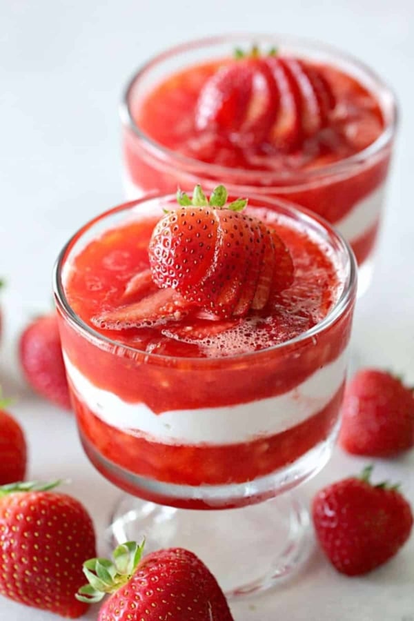 Two glass cups filled with layered strawberry and cream dessert, topped with sliced strawberries, surrounded by whole strawberries on a white surface.