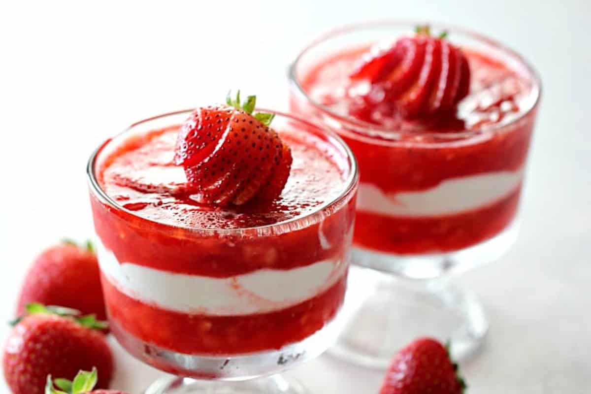 Two glass cups of strawberry parfait with layered strawberries and cream, each topped with a sliced strawberry; whole strawberries are placed nearby.