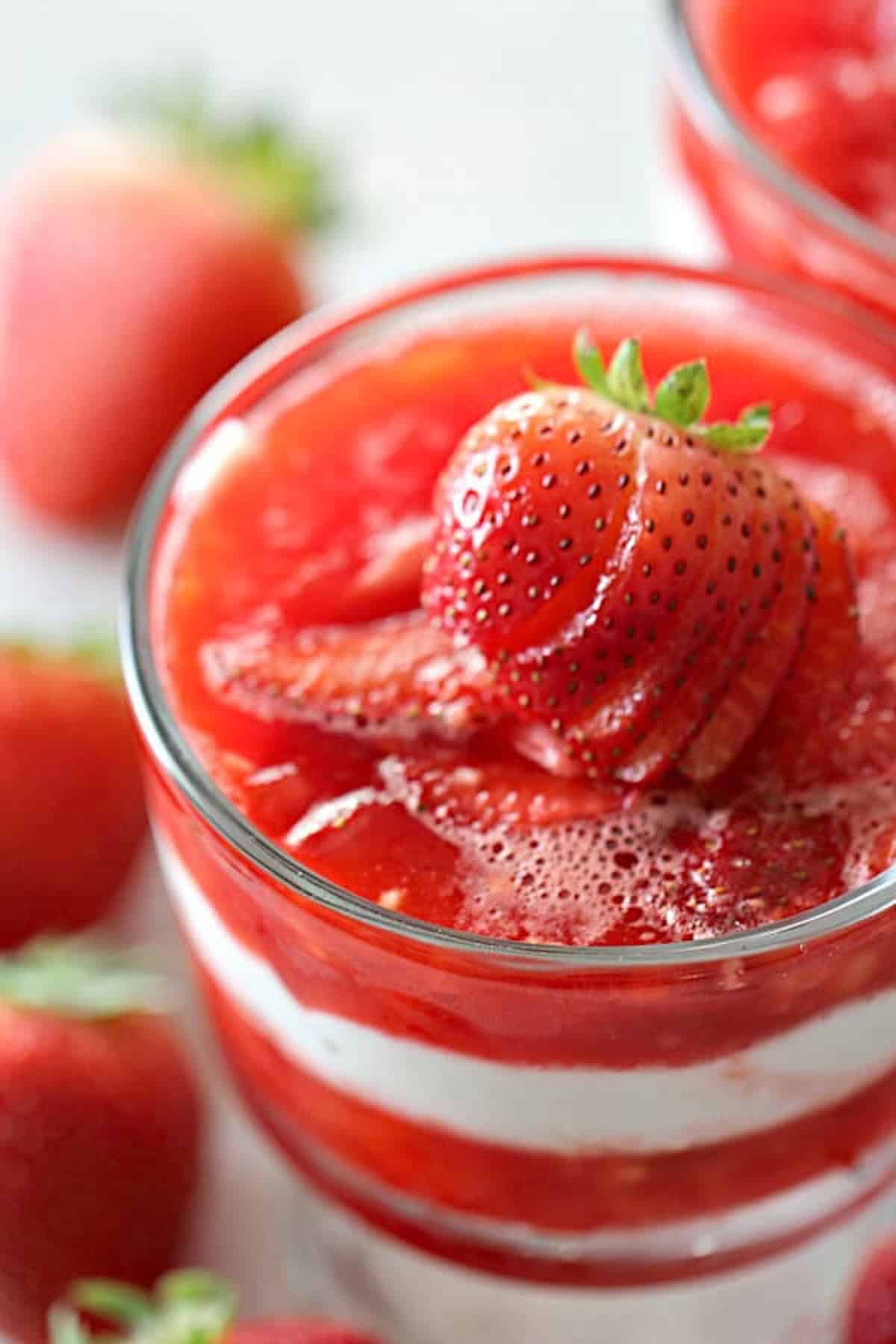 A glass filled with layers of strawberry sauce and cream, topped with sliced fresh strawberries; whole strawberries are visible in the background.