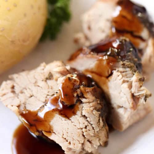 Slices of cooked pork loin with brown gravy on a white plate, served with a boiled potato and green vegetable garnish in the background.