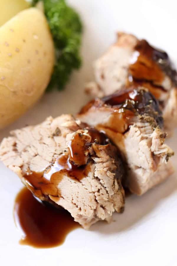 Slices of cooked pork loin with brown gravy on a white plate, served with a boiled potato and green vegetable garnish in the background.