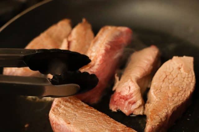 Tongs are holding a piece of meat while several other pieces are cooking in a frying pan.
