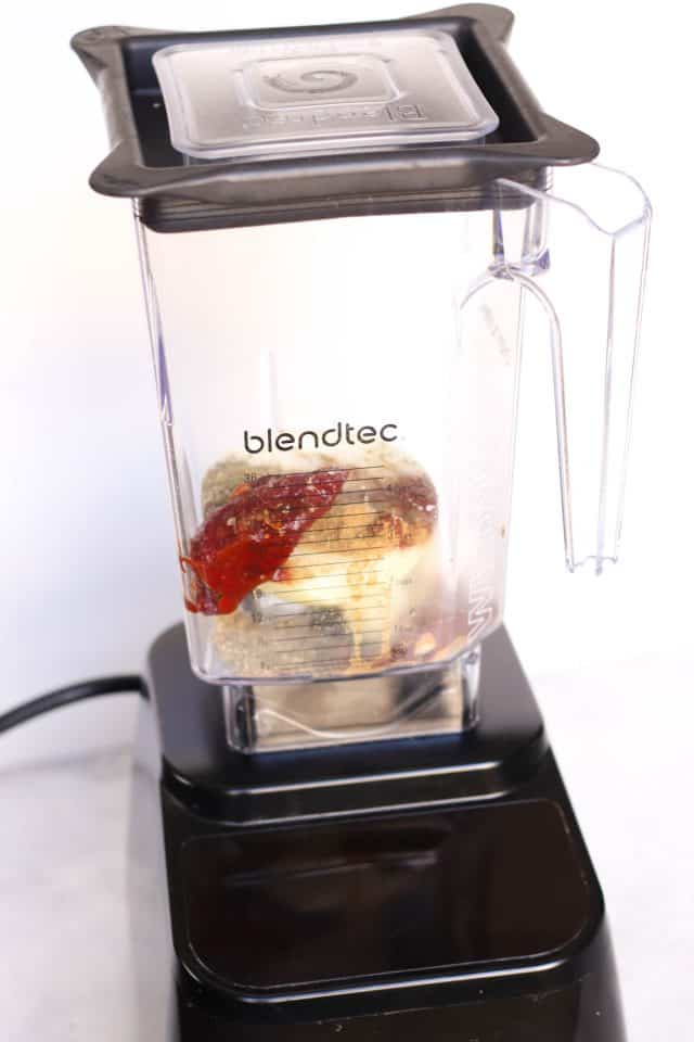 A Blendtec blender with a transparent jar containing ingredients, including a circular object with layered sections and what appears to be a reddish sauce. The blender is on a white surface.